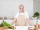 A mature woman in an apron stands at a kitchen counter with fresh produce and cheese, ready for cooking.